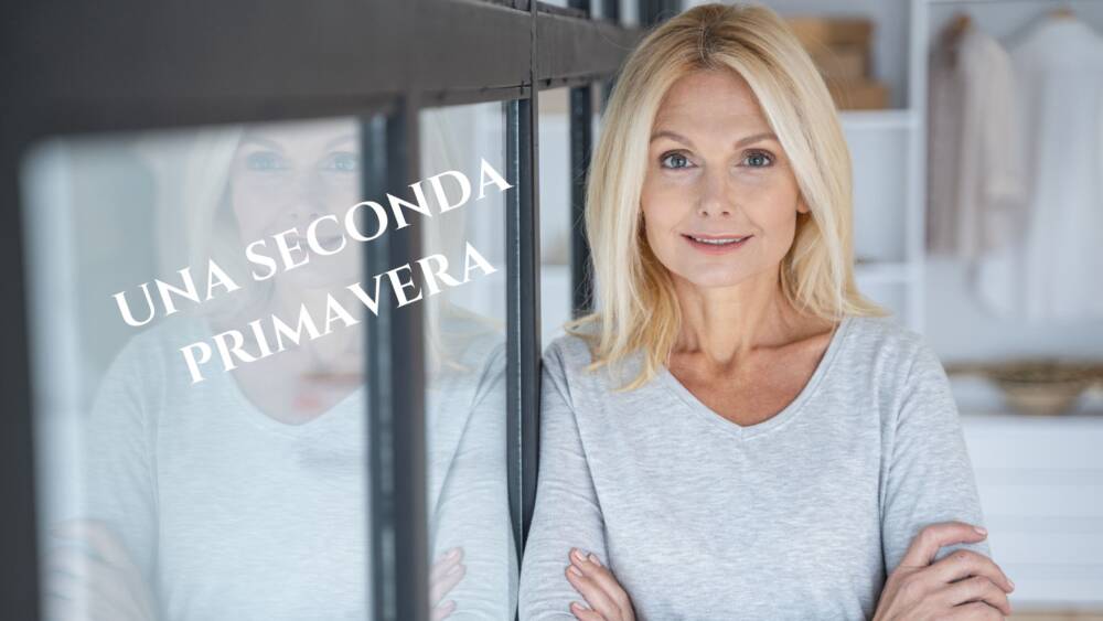 Una donna bionda di mezza etá, sorridente, con una maglietta bianca, appoggiata a una finestra
